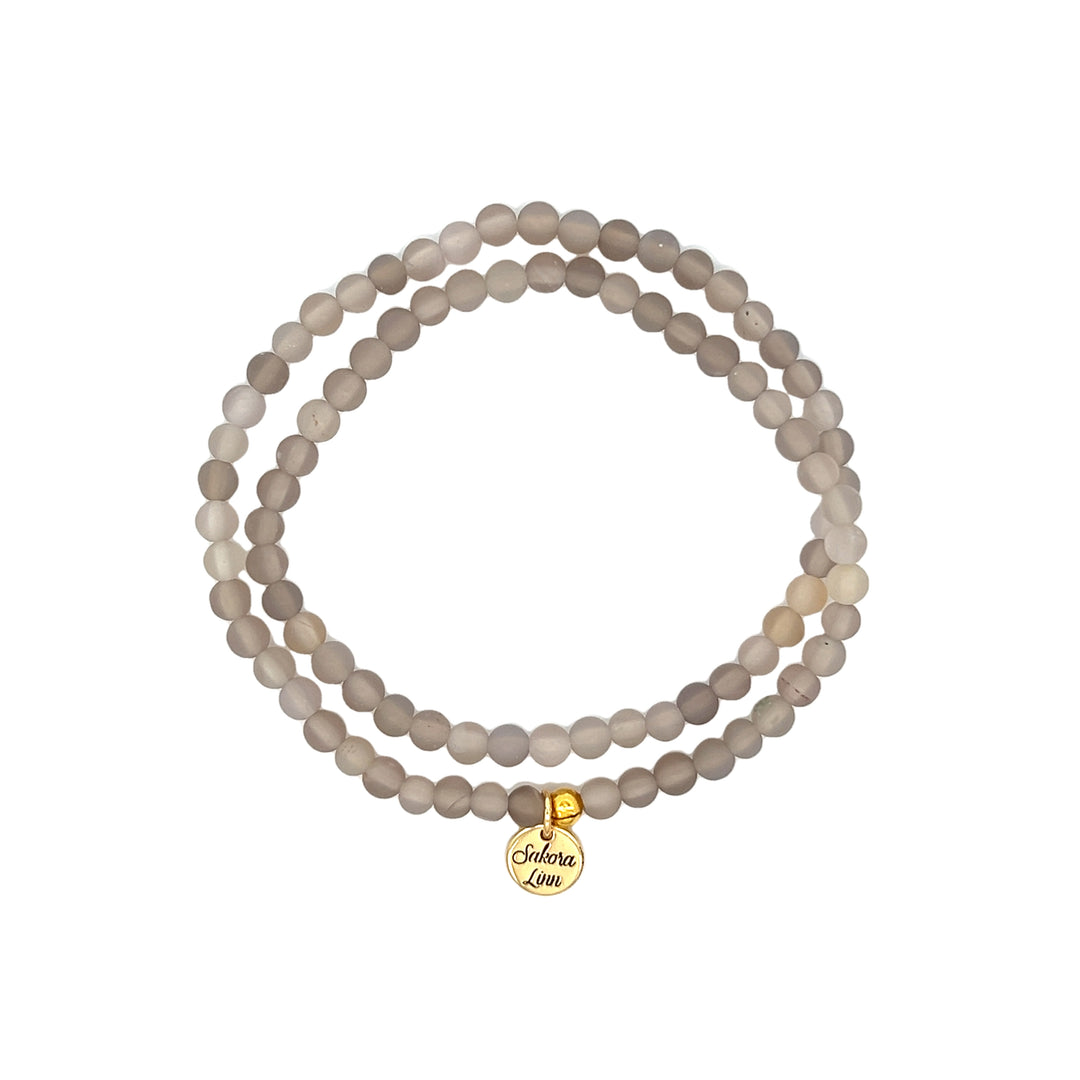 Close-up of an agate stretch style double wrap bracelet made with 4mm frosted light Gray Agate Beads, a Clear Cord, 18k Gold Plated Brass Logo Tag, displayed on a white background.