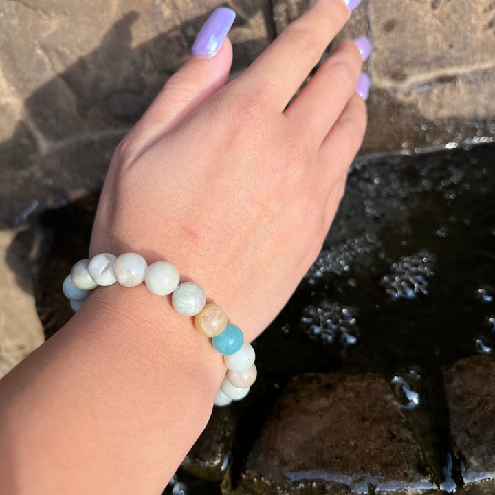 A closeup of a woman's hand and wrist with lavander nail polish, wearing a 10mm amzonite stretch style bracelet, with a blurred brown and gray rock with a pool of water in the background.