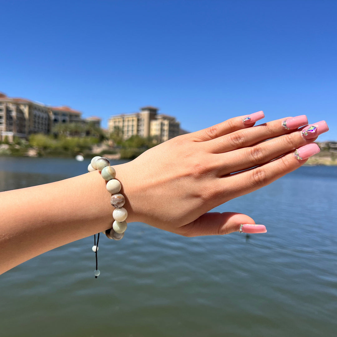Woman's wrist and hand with pink fingernails, wearing a 12mm Amazonite pull-tie bracelet with shades of pale blue, green and white, with a lake and blurred buildings in the background.