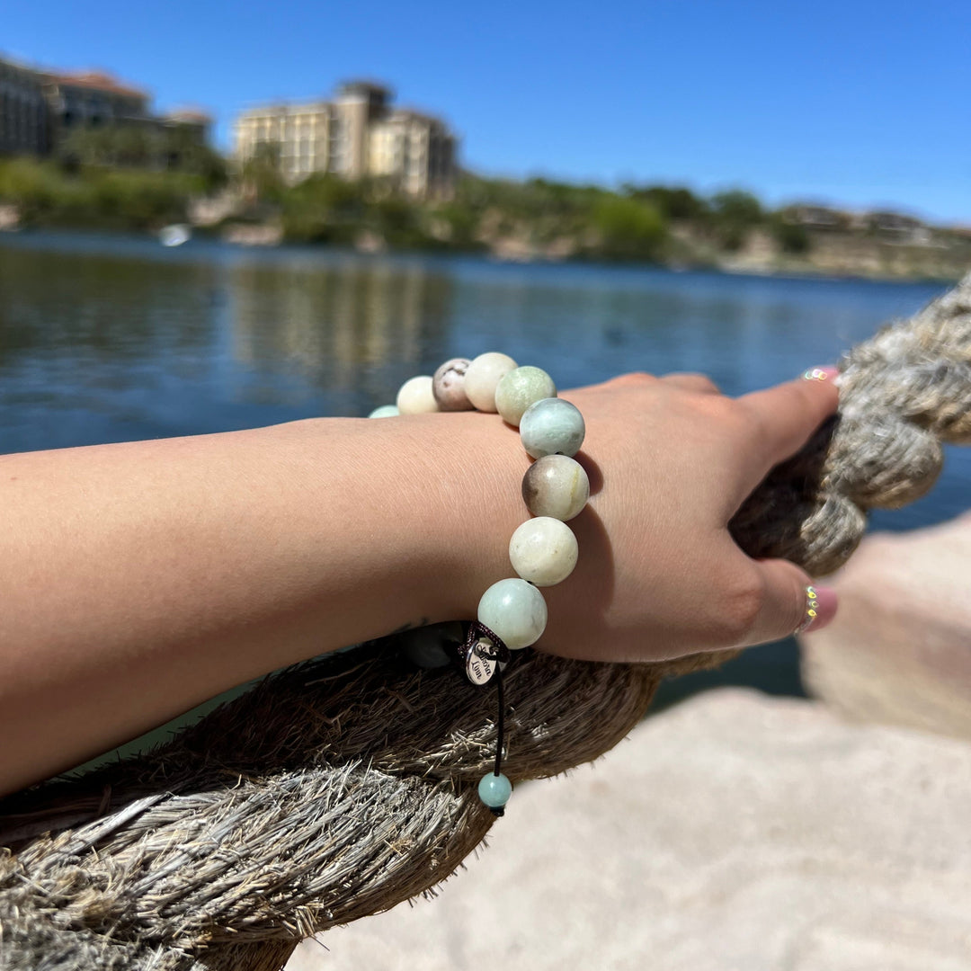 Woman's wrist and hand laying on top of a rope, wearing a 12mm Amazonite pull-tie bracelet in shades of pale blue and green, with a silver sakora linn logo charm, with a lake and blurred buildings in the background.