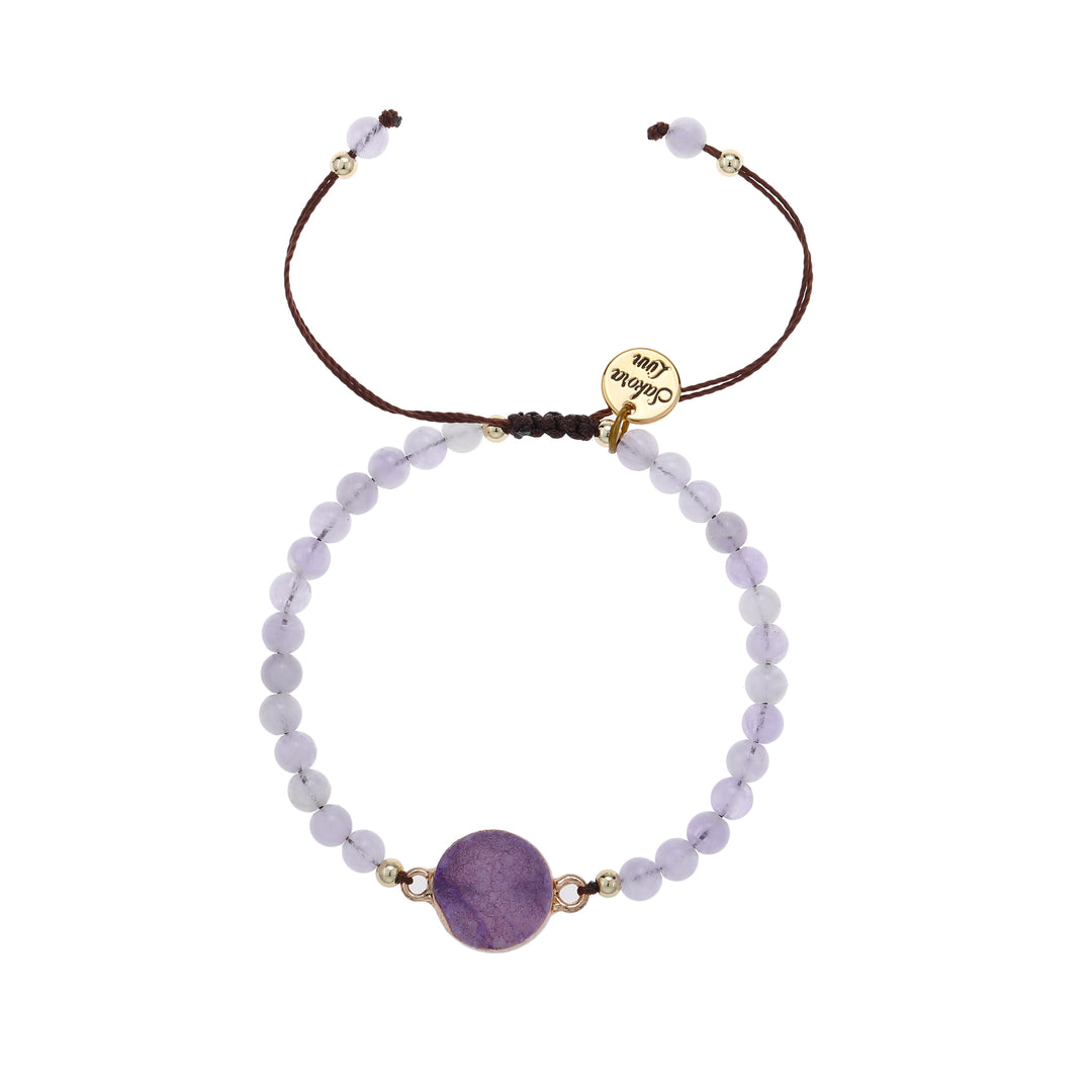 Close-up of an adjustable slip knot style bracelet with 4mm round lavender beads, a 10mm purple amethyst druzy stone, mounted in a gold setting, in the center of the bracelet, with small gold accent, strung on a dark brown cord with a round gold logo charm stamed with Sakora Linn in black, displayed on a white background.