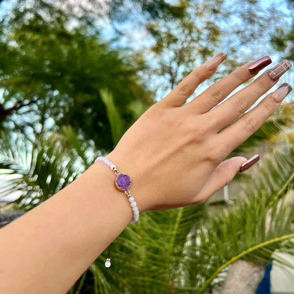 Close-up of a woman's hand with long taupe and mauve fingernails, wearing a purple and lavender color amethyst druzy stone bracelet, with blurred green palm trees and blue sky in the background.