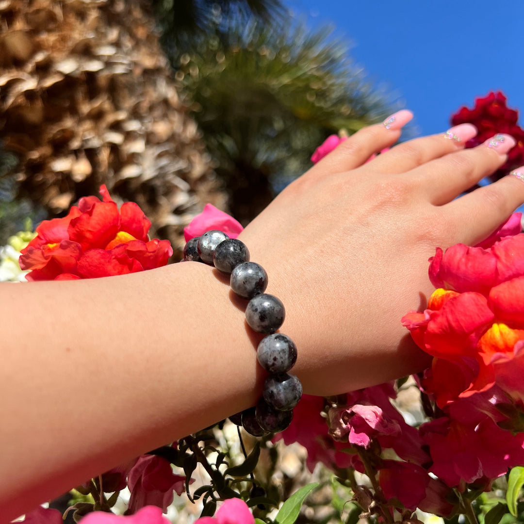 Woman's wrist and hand with pink fingernails, wearing a 12mm black labradorite pull-tie bracelet, with red and pink flowers and a blurred palm tree behind the hand.