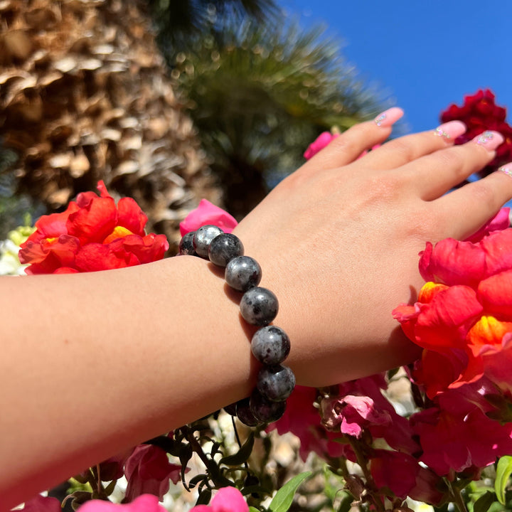 Woman's wrist and hand with pink fingernails, wearing a 12mm black labradorite pull-tie bracelet, with red and pink flowers and a blurred palm tree behind the hand.