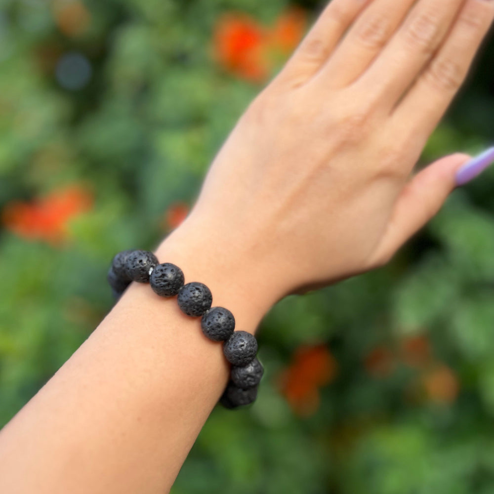 Close-up of a woman's wrist wearing a black lava stone stretch style bracelet made with round 12mm beads, with blurred background of green plants and orange flowers.