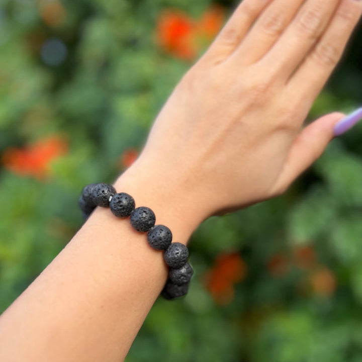 Close-up of a woman's wrist wearing a black lava stone stretch style bracelet made with round 12mm beads, with blurred background of green plants and orange flowers.