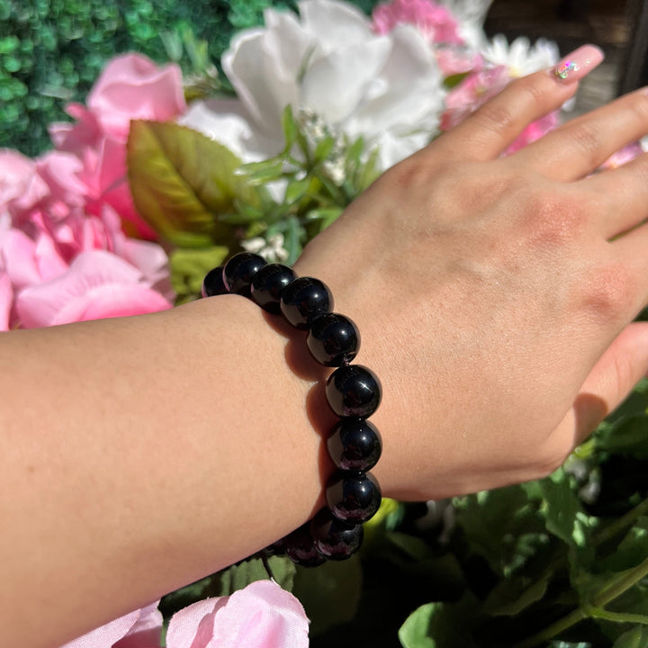 A close-up of a woman's wrist wearing a 12mm glossy blck onyx bracelet with blurry pink, white, and green flowers in the background.