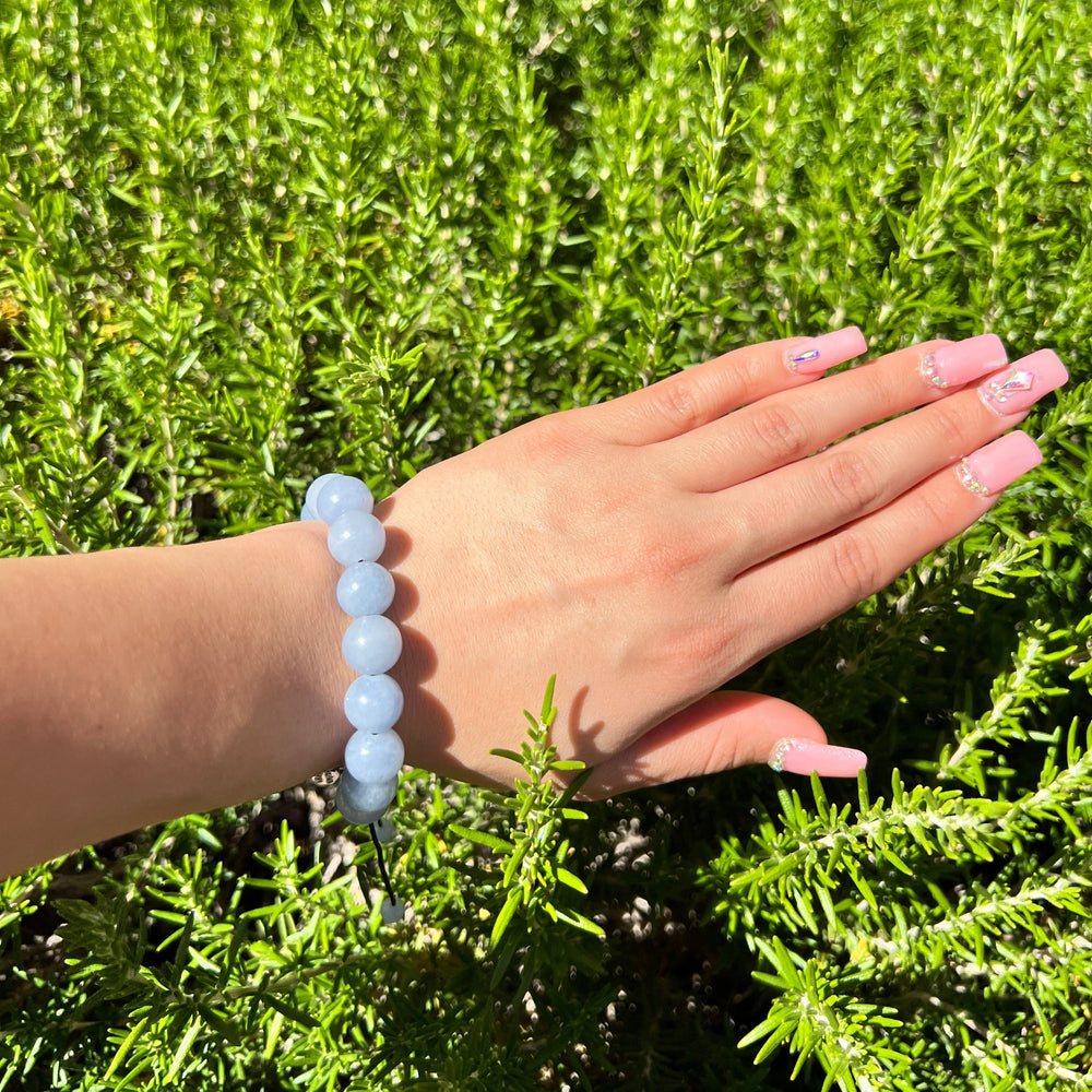 Close-up of a woman's hand and wrist with pink fingernails wearing a 12mm blue aquamarine bead bracelet with a green plant background.