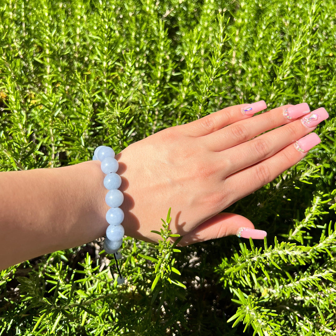 Close-up of a woman's hand and wrist with pink fingernails wearing a 12mm blue aquamarine bead bracelet with a green plant background.