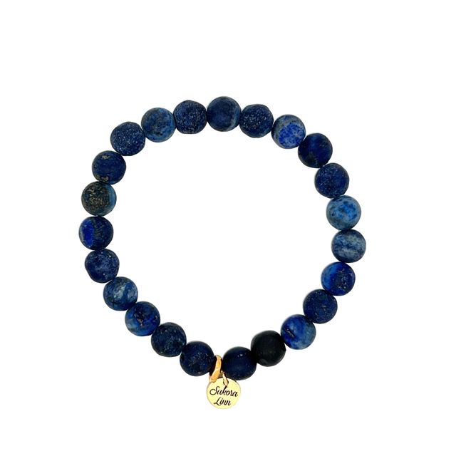Close-up of a stretch style blue lapis bracelet made with 8mm round matte blue lapis beads, in shades of dark blue and medium blue, with one black clay bead, an 18k gold plated logo tag engraved with Sakora Linn, displayed on a white 
background.