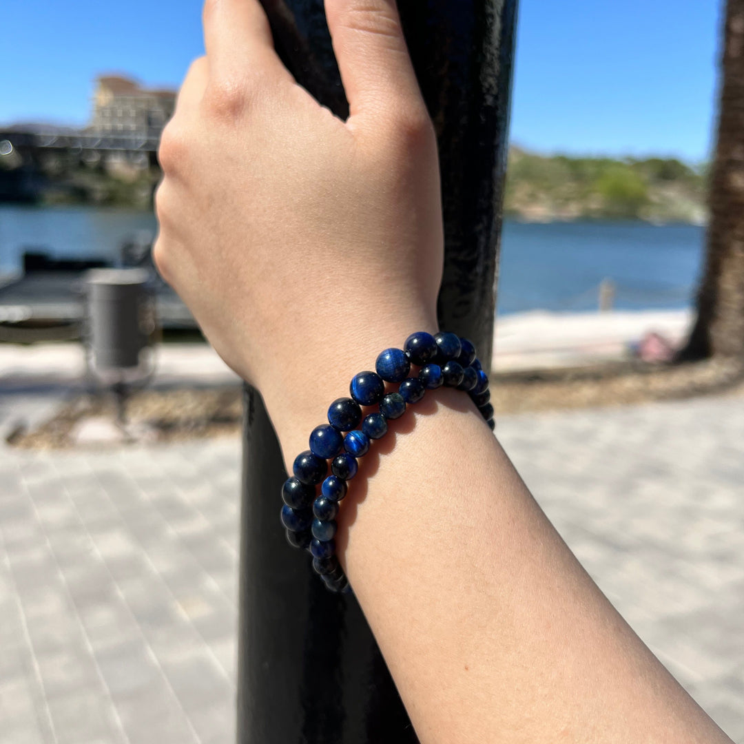 A close-up of a hand and wrist, outside, holding on to a pole, wearing two stretch bracelets with round blue tigers eye beads, one with 6mm beads, and one with 8mm beads, with a blured background of water, a building, a green hill, and gray pavers in the foreground.