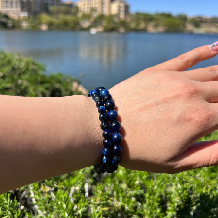 A close-up of a hand and wrist, outside, wearing two stretch bracelets with round blue tigers eye beads, one with 6mm beads, and one with 8mm beads, with a blured background of water, buildings, and green plants.