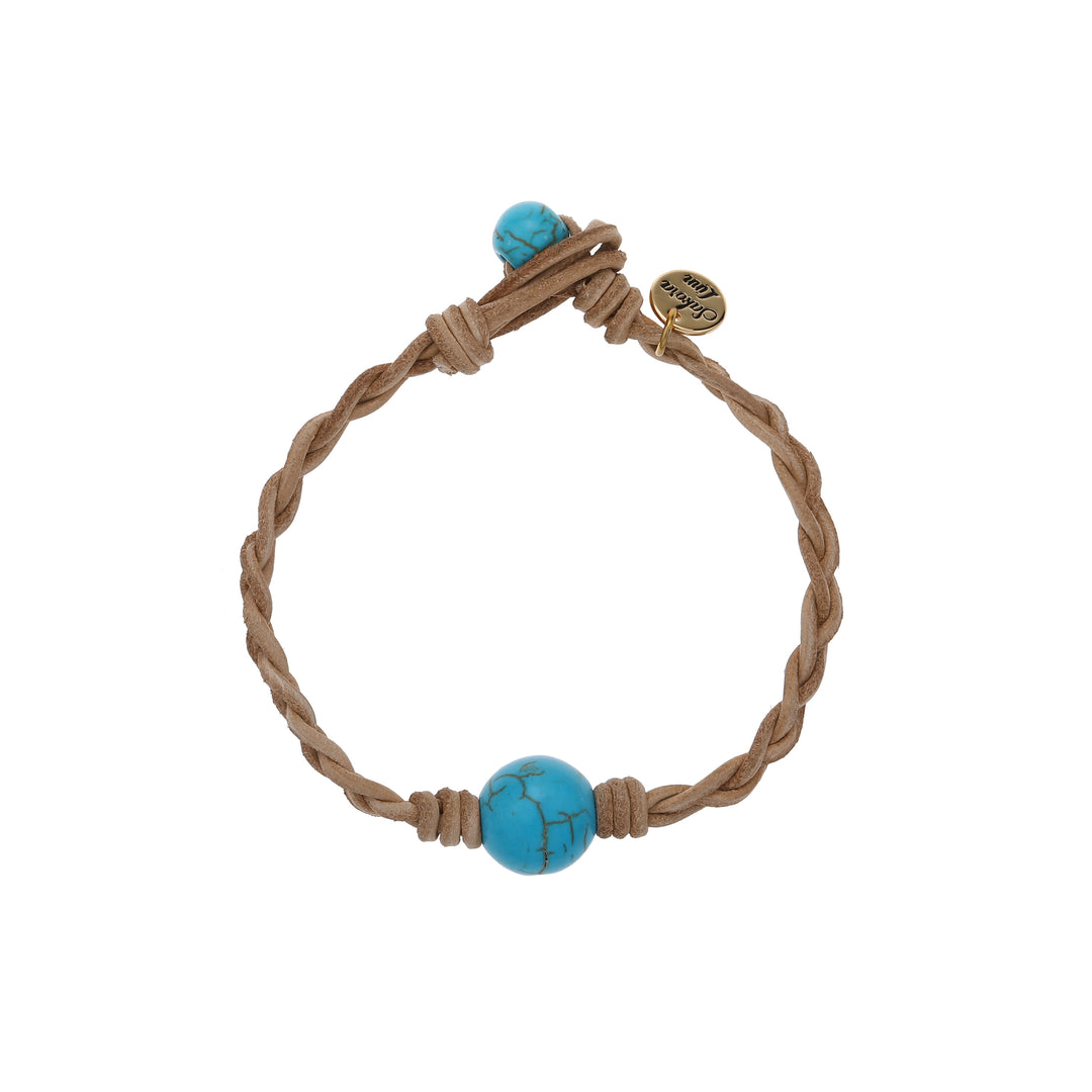 Close-up of a braided bracelet made with ltan leather cord, a blue cracked turquoise center bead, and a smaller turquoise bead for the toggle style closure, and a gold logo cahrm stamped with Sakora Linn, displayed on a white background.