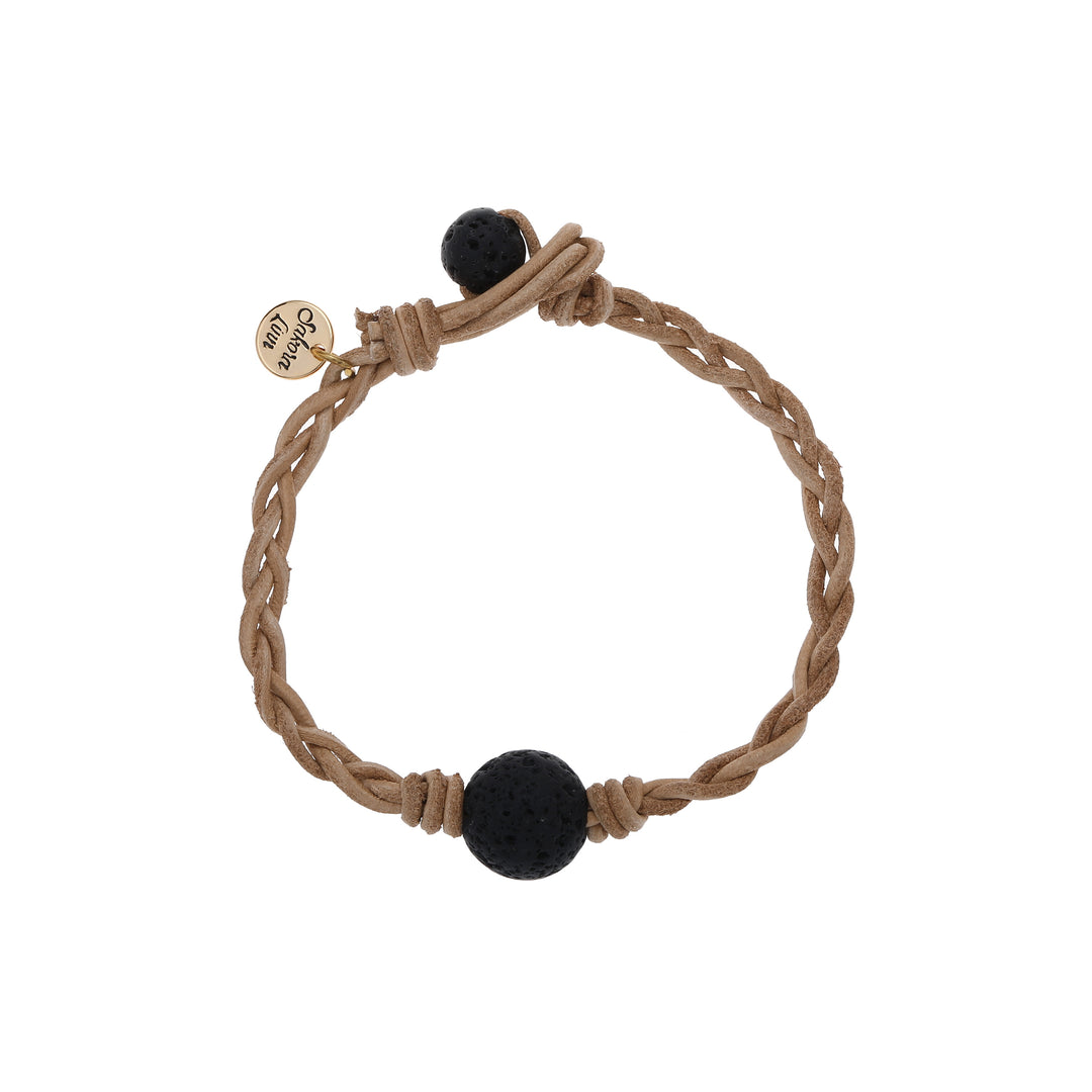 Close-up of a braided bracelet made with ltan leather cord, a black lava stone center bead, and a smaller blaclk lava stone bead for the toggle style closure, and a gold logo cahrm stamped with Sakora Linn, displayed on a white background.
