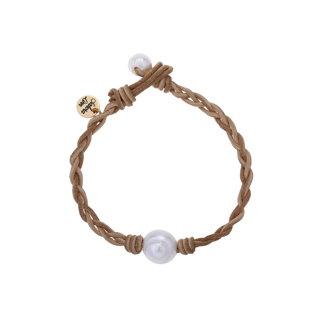 Close-up of a braided bracelet made with ltan leather cord, a white fresh water pearl center bead, and a smaller pearl bead for the toggle style closure, and a gold logo cahrm stamped with Sakora Linn, displayed on a white background.