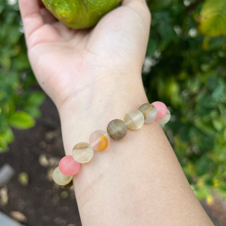 A woman's wrist wearing a cherry quartz stretch style bracelet made with 10mm stones in shades of pink, tan, white, and gray, with a blurred background of green plants.