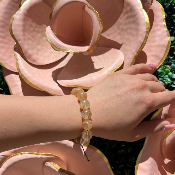 A close-up of a woman's hand and wrist wearing a pale yellow citrin 12mm bead bracelet with a large metal pale pink flower sculpture in the background.