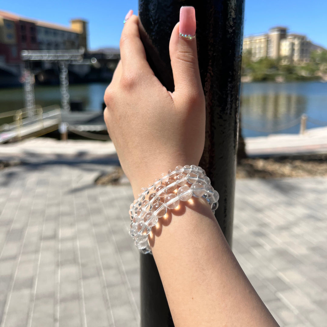 A close-up of a hand and wrist with pink fingernails, outside holding on to a pole, wearing three stretch bracelets with round clear quartz beads, one with 6mm beads, one with 8mm beads, and one with 10mm beads, with a blured background of water, a building, a green hill, and gray pavers in the foreground.