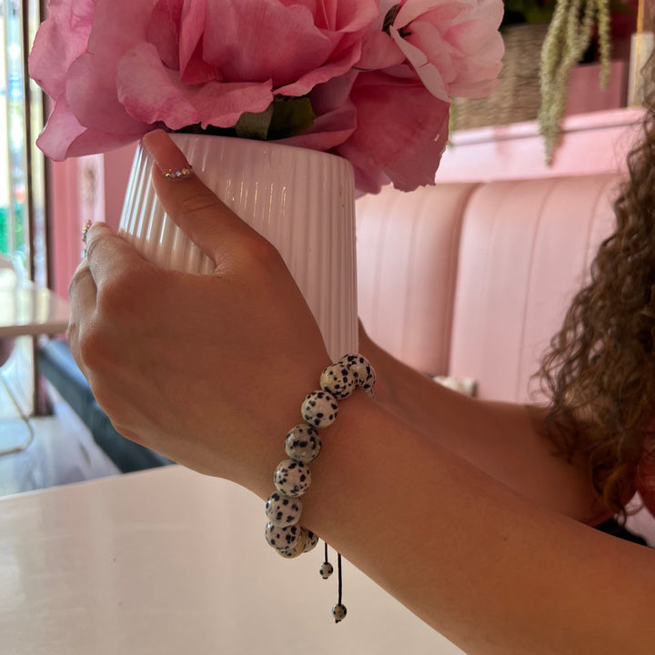 A woman's hands and wrist holding a pot of flowers wearing a 12mm black and white Dalmatian stone bracelet, with a blurry pink booth and white table in the background.