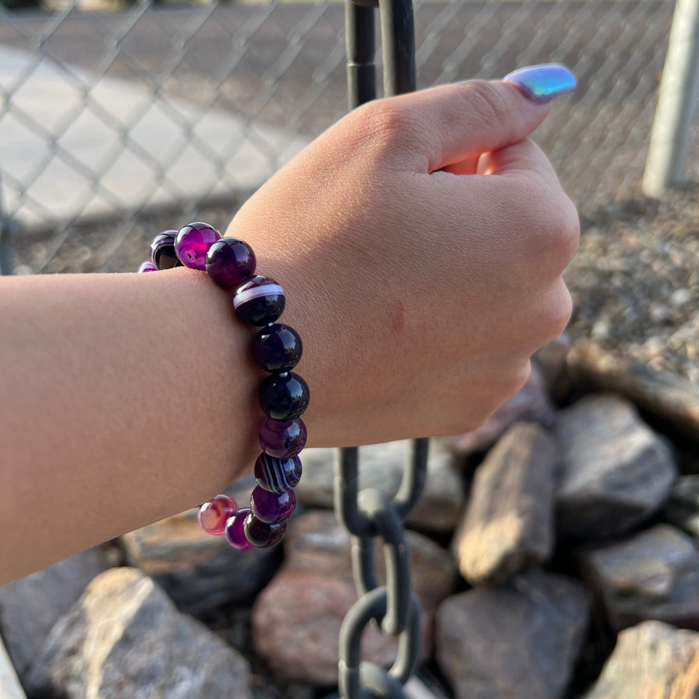A woman's wrist wearing a 10mm dog teeth amethyst stretch style beaded bracelet, with blurred gray rocks and a black chain in the background.