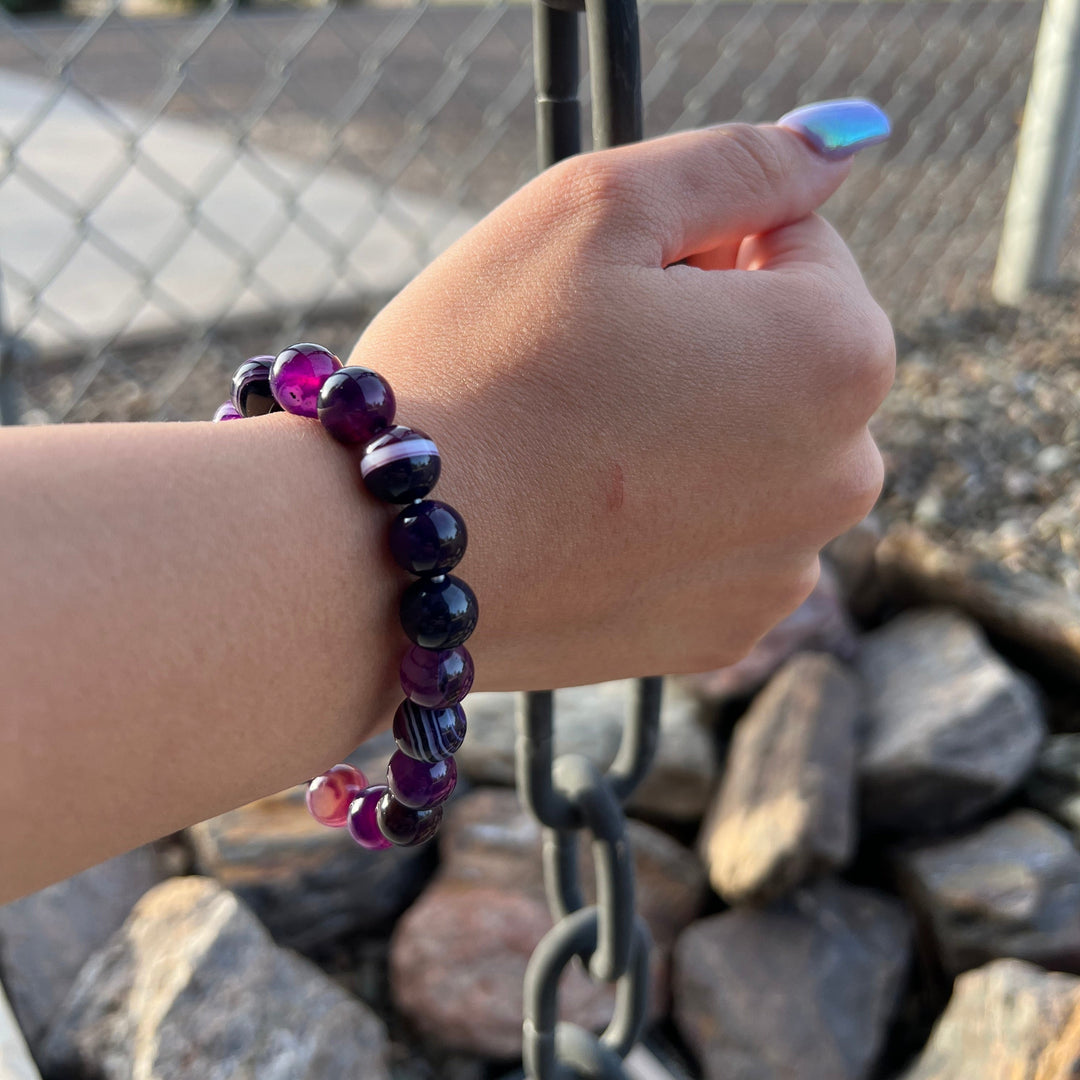 A woman's wrist wearing a 10mm dog teeth amethyst stretch style beaded bracelet, with blurred gray rocks and a black chain in the background.