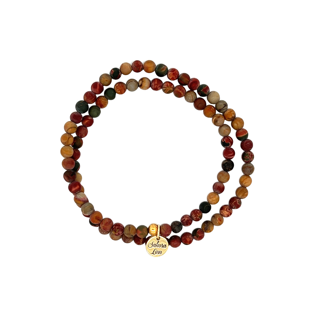 Close-up of a double wrap stretch style bracelet made with 4mm jasper beads, in shades of deep red, tan, and black; with an 18k gold plated logo tag engraved with Sakora Linn, displayed on a white background.