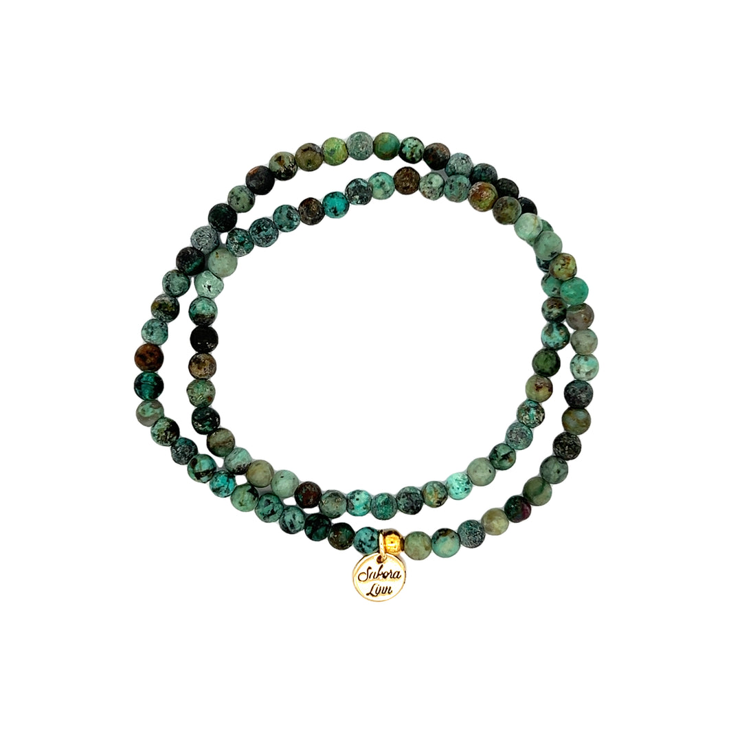 Close-up of a double wrap stretch style bracelet made with 4mm matte turquoise beads, in shades of green and black, with an 18k gold plated logo tag engraved with Sakora Linn, displayed on a white background.