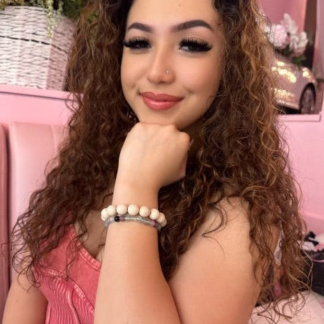 Close-up of a woman with long curly brown hair smiling wearing a tan fossil stone bracelet and a flourite bracelet on her wrist, with a pink background behind her.