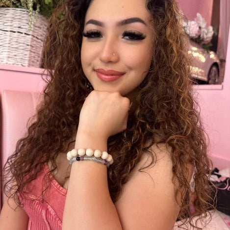 A close-up of a woman with long brown curly hair smiling, wearing a pink top and sitting in a pink booth, with her hand under her chin wearing a creamy white 12mm bead fossil stone bracelet.