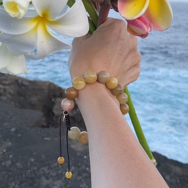 A woman's wrist and hand holding a bouquet of Hawaiian Plumeria flowers wearing a golden jade bracelet with 12mm yellow beads and pull tie closure with gold logo charm, and a blurry background of gray rocks and blue ocean.