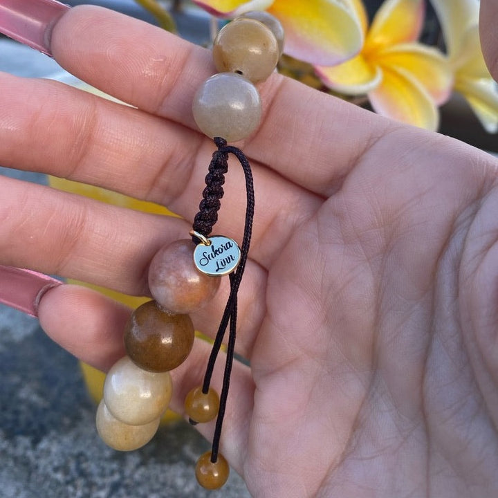 A woman's hand holding a yellow golden jade pull tie bracelet made with 12mm beads, showing a gold logo charm with sakora linn engraved on it, with blurry yellow plumaria flowers and gray rock in the background.
