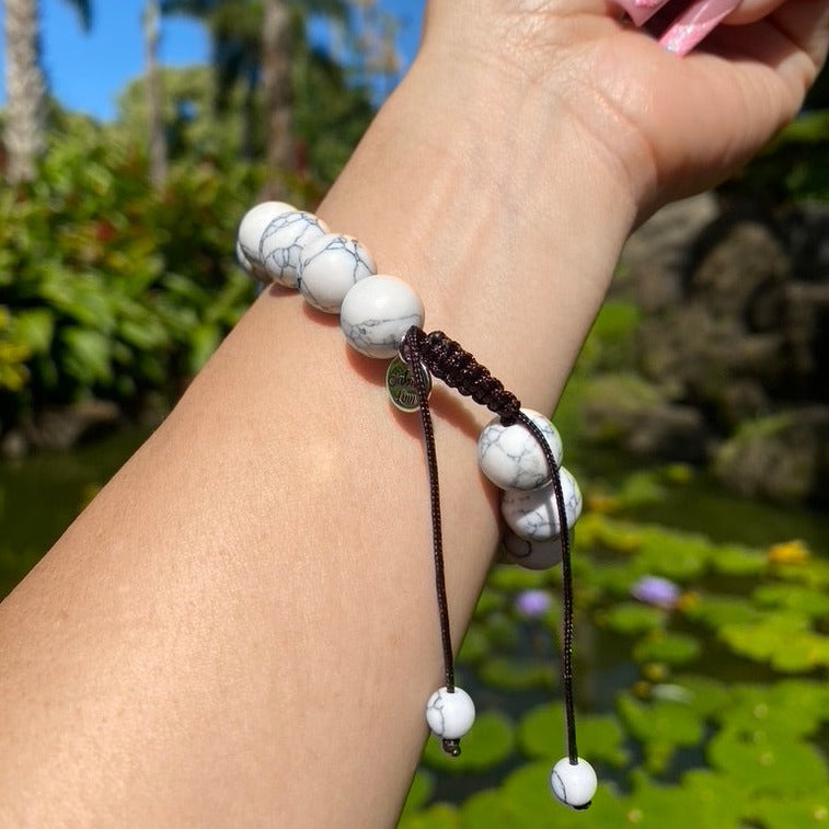 A close-up of the underside of a woman's wrist wearing a white marbled 12mm braclet with a dark brown pull tie closure and a silver logo charm stamped with Sakora Linn, with a blurry lily pond and green trees in the background.