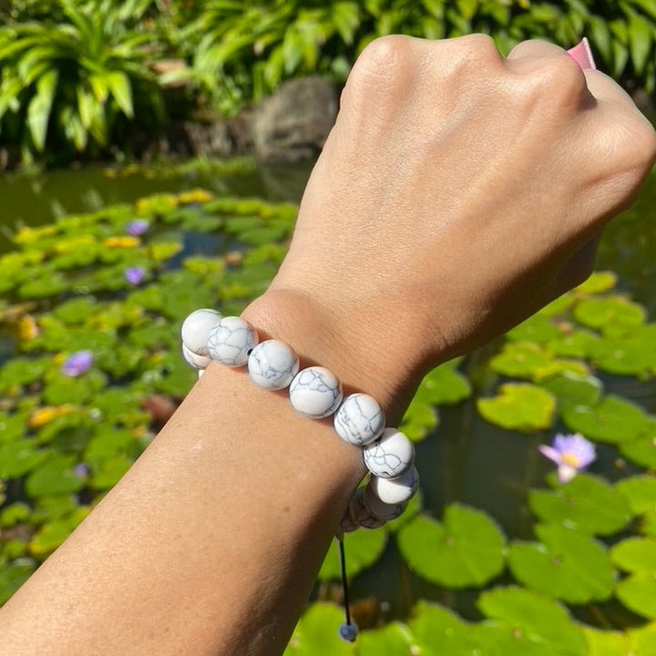 A close-up of the top of a woman's wrist wearing a white marbled 12mm braclet with a blurry green lily pond and green trees in the background.