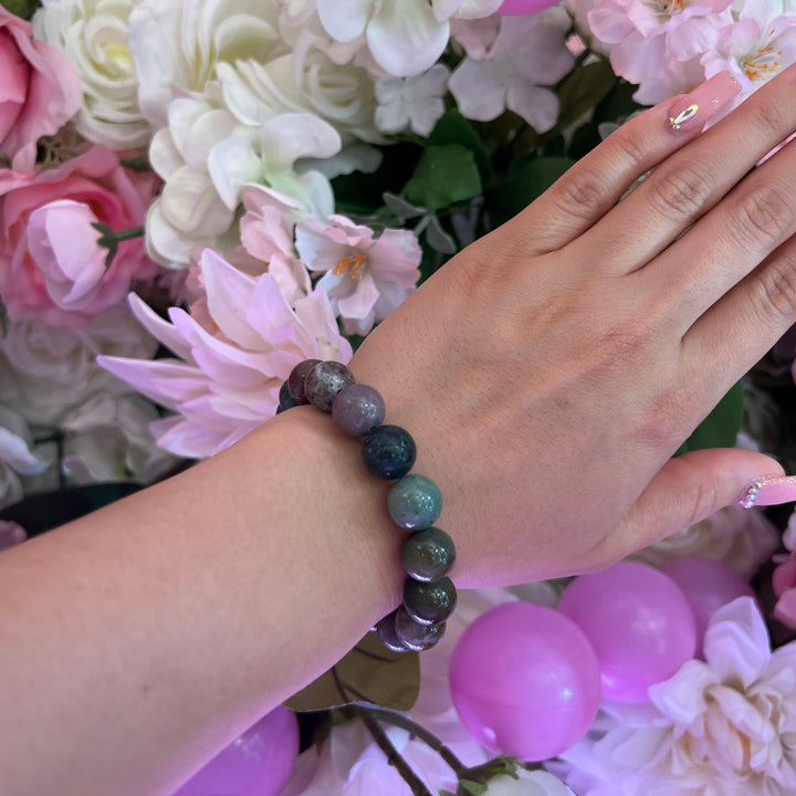 Close-up of a woman's wrist wearing an Indian agate bracelet with shades of green and purple 12mm round stone beads, with pink and white flowers in the background.