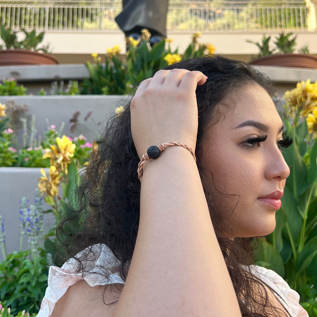 Close-up of the side of a woman's face with long black hair, holding her hand on her hair, wearing a braided leather lava stone bracelet, with a building, green plants, and yellow flowers in the background.