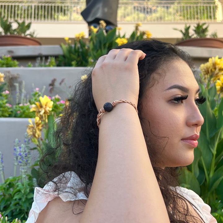 Close-up of the side of a woman's face with long black hair, holding her hand on her hair, wearing a braided leather lava stone bracelet, with a building, green plants, and yellow flowers in the background.