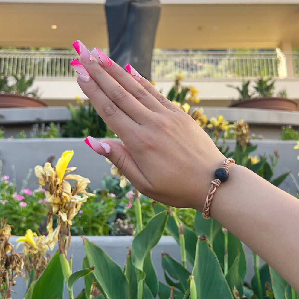 A close-up of a woman's hand with long pink fingernails wearing a braided leather bracelet with a lava stone bead, with a building, green plants, and yellow flowers in the background.