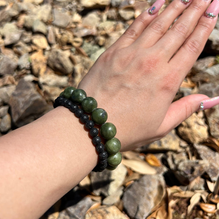 Close-up of a woman's wrist wearing a 6mm black lava stone stretch style bracelet and a 12mm olive jade stretch bracelet, with blurred background of tan and gray rocks.