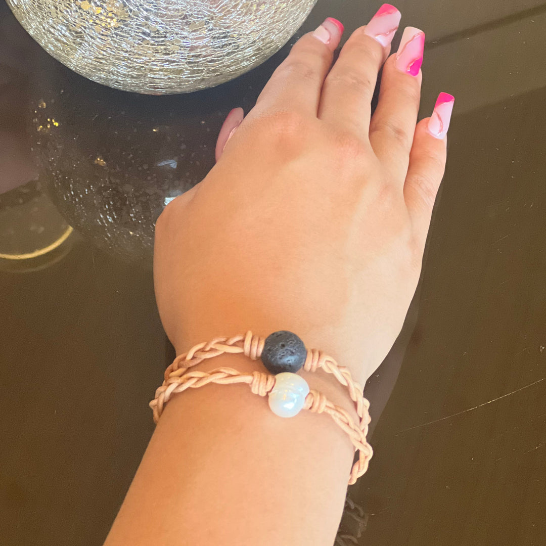 A close-up of a woman's hand resting on a shiny brown table, wearing two braided leather bracelets, one with a lava stone and one with a pearl in the center of the bracelet.