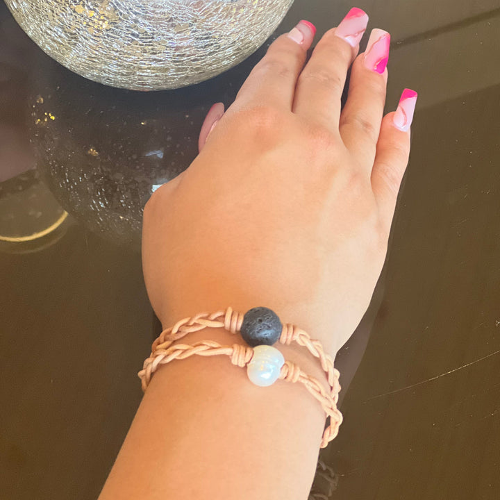 A close-up of a woman's hand resting on a shiny brown table, wearing two braided leather bracelets, one with a lava stone and one with a pearl in the center of the bracelet.