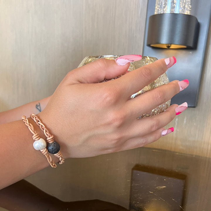 A close-up of a woman's hand with pink fingernails holding a gold glass ball, wearing two leather braided bracelets, one with a lava stone bead and one with a pearl bead, with tan wall and black light sconce in the background.