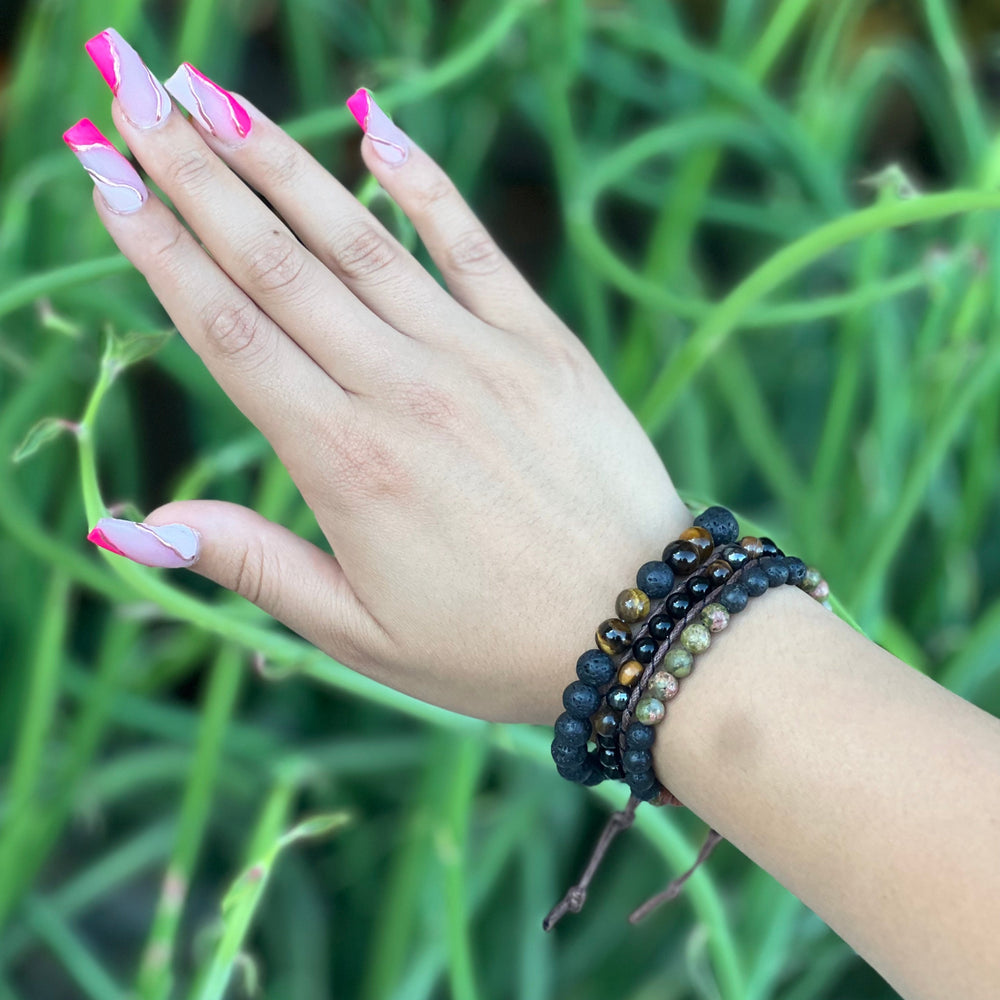 Close-up of a woman's wrist with pink nail polish, wearing three bracelets, one is a stretch style lava stone and tiger's eye bracelet made with 8mm round black lava stone beads with four 8mm tiger's eye beads on each side of one black center bead, in black and brown colors, one is a unakite and lava stone 6mm bead bracelet in black and shades f green, and a wrap bracelet with 6mm tiger's eye, hematite and onyx beads in black and brown colors, with a blurred green plant background.