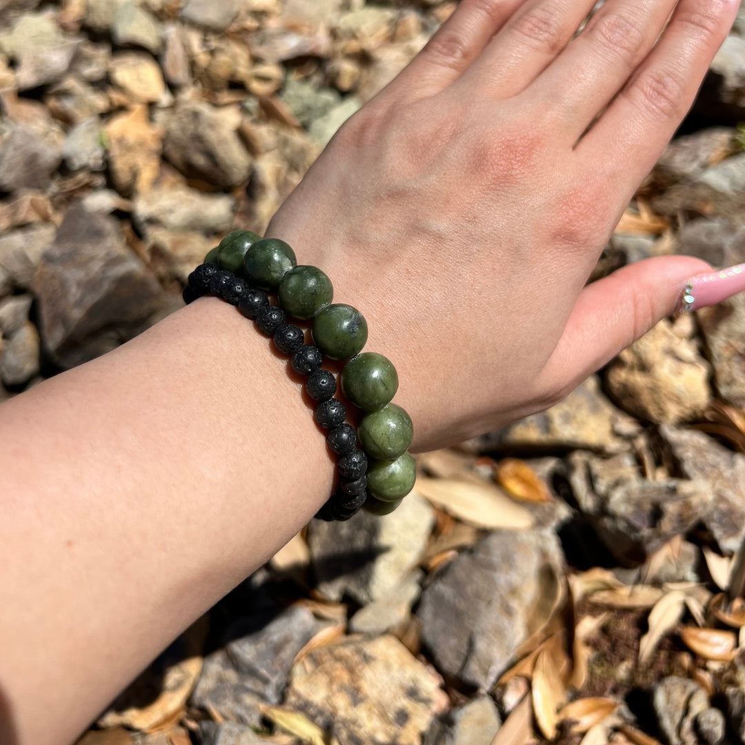 Close-up of a woman's wrist wearing a 12mm green olive jade bracelet and an 8mm lava stone bracelet, with blurry tan rocks in the background.