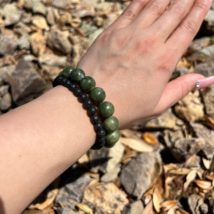 Close-up of a woman's wrist wearing a 12mm green olive jade bracelet and an 8mm lava stone bracelet, with blurry tan rocks in the background.