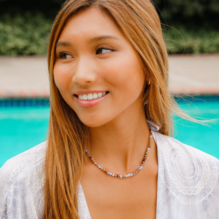 Close-up of a teen aged girl smiling with golden dark blonde hair wearing a multicolor beaded necklace, with a blurred swimming pool in background. 
