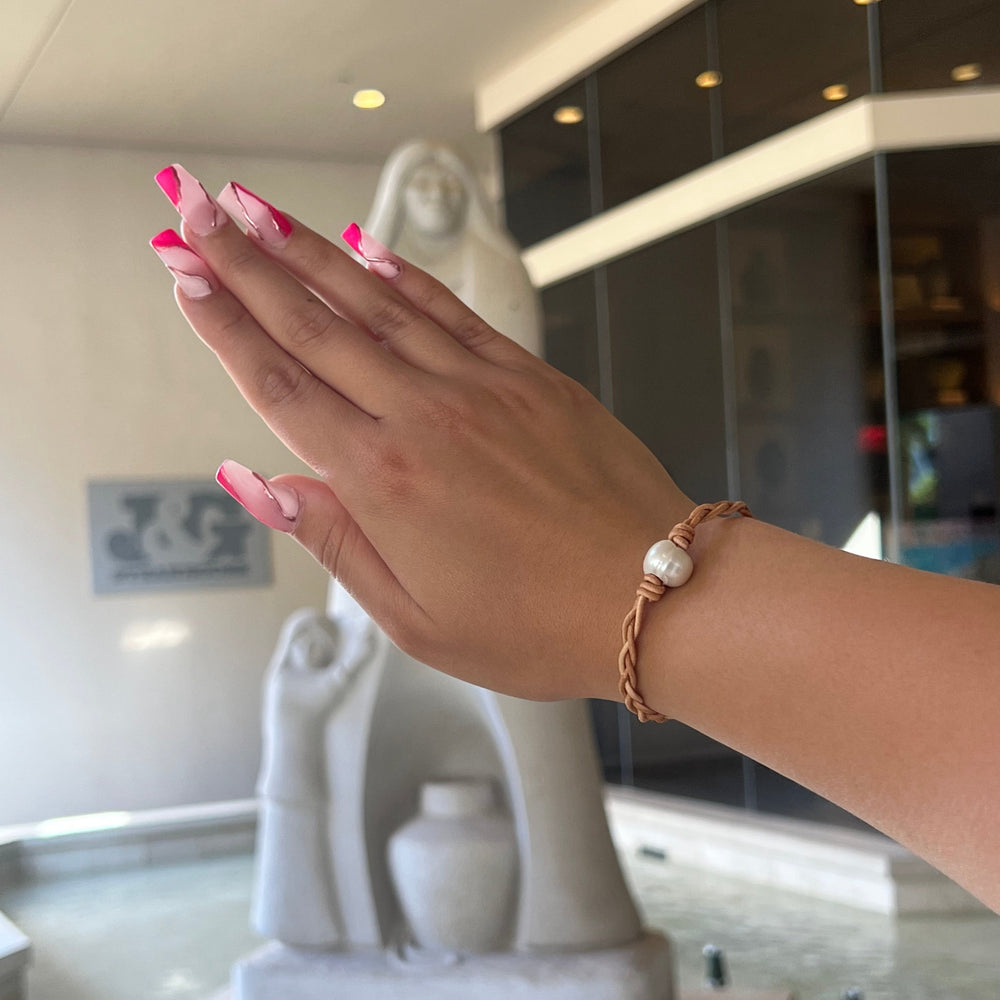 A woman's hand with pink fingernails wearing a tan braided leather with a center white pearl, with a blurred white sculpture and glass windows in a building lobby in the background.