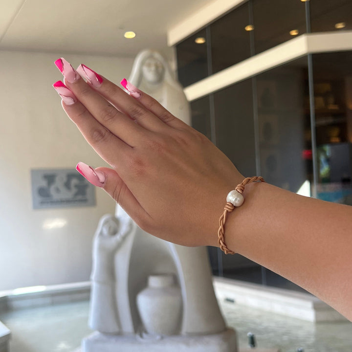 A woman's hand with pink fingernails wearing a tan braided leather with a center white pearl, with a blurred white sculpture and glass windows in a building lobby in the background.