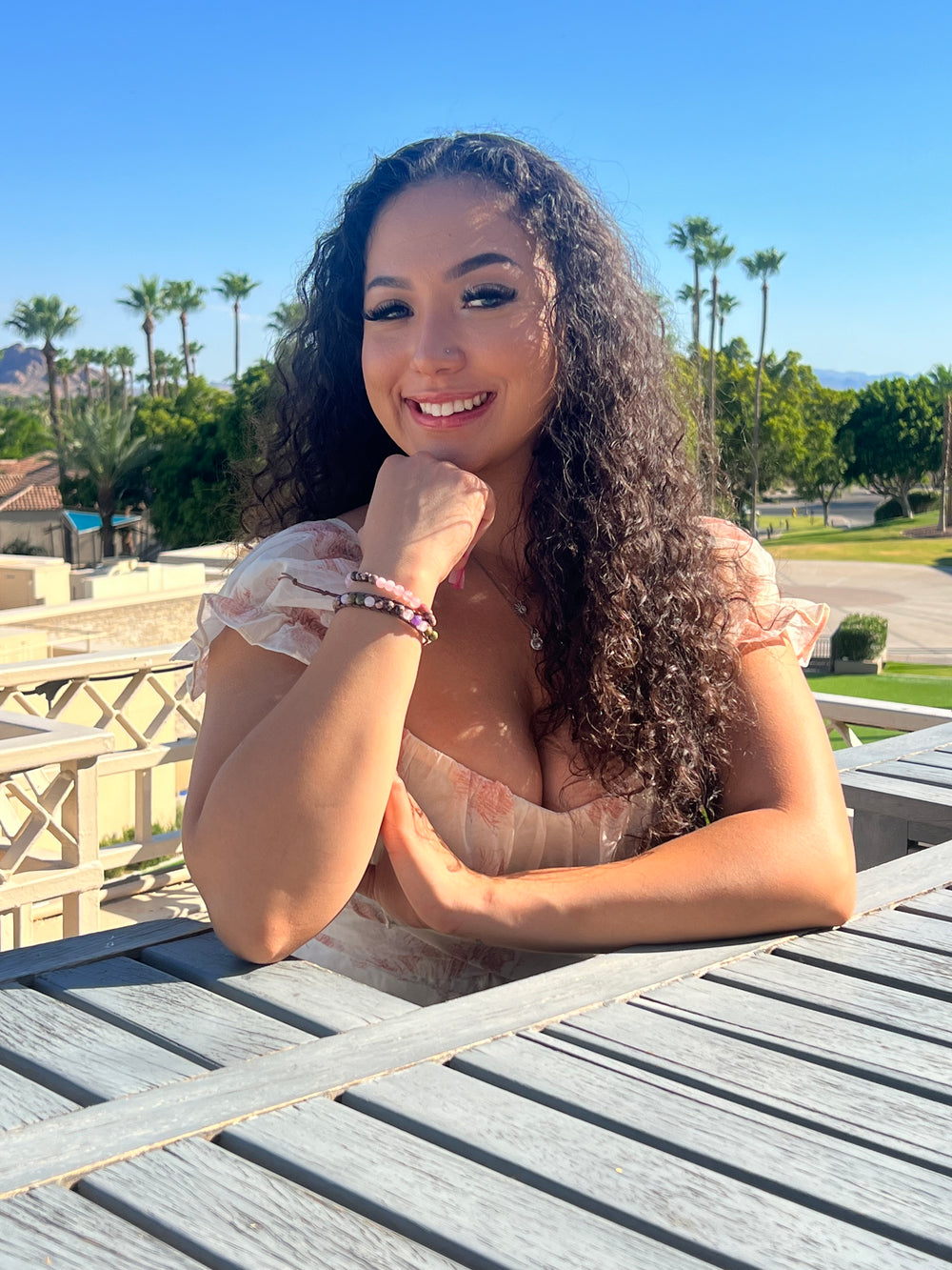 A woman with long dark curly hair smiling with her hand under her chin, wearing two bead bracelets in shades of pink and a white dress with pink flower print, sitting at a table on a balcony, with a park and green trees in the ackground.