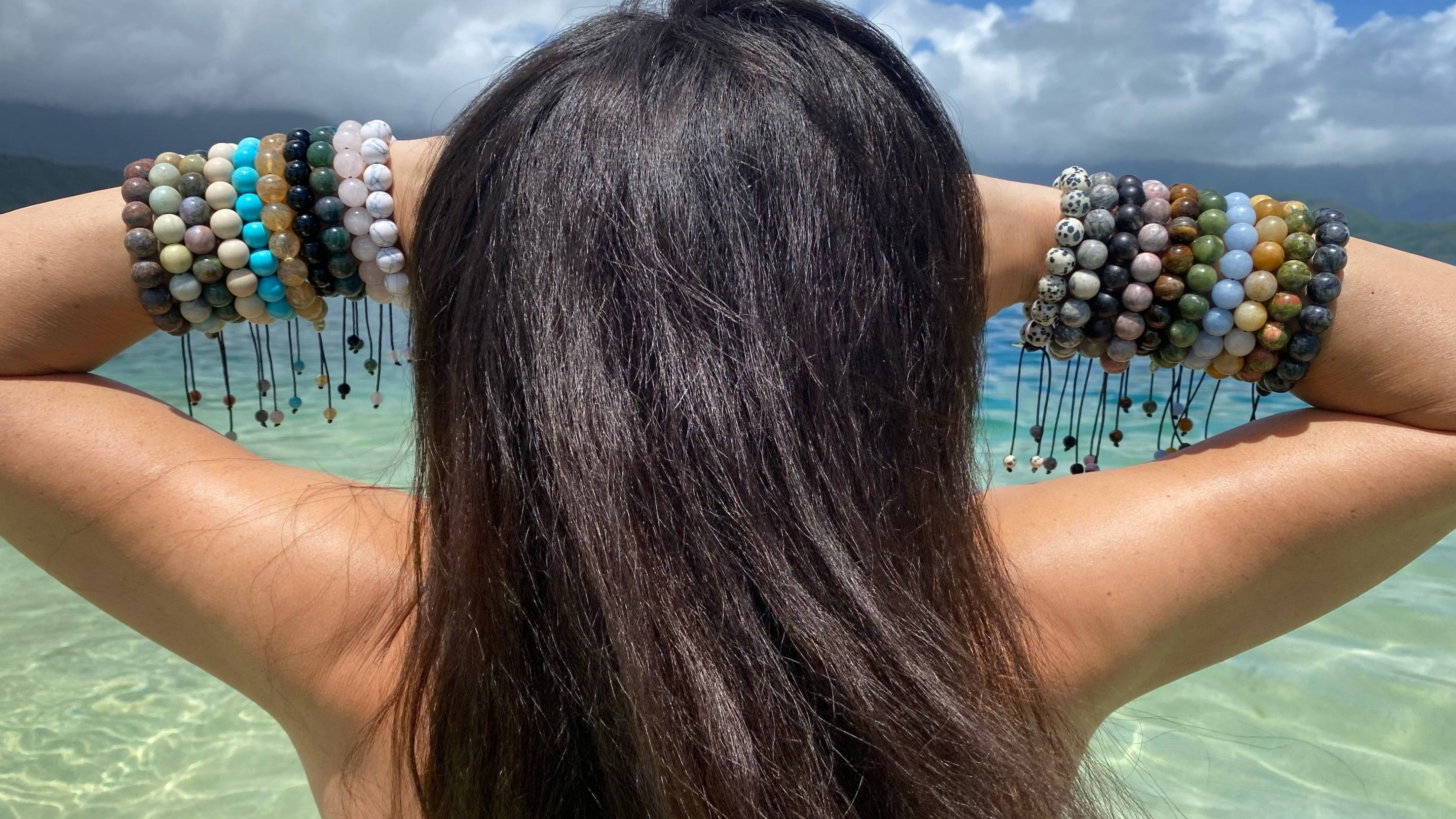 Close-up of the back of a woman with long black hair with ten large bead bracelets on each wrist and holding her hands to the front of her face with arms bent outwards, in the background there's a blue sky, stormy clouds, and the ocean water in shades of blue and green.