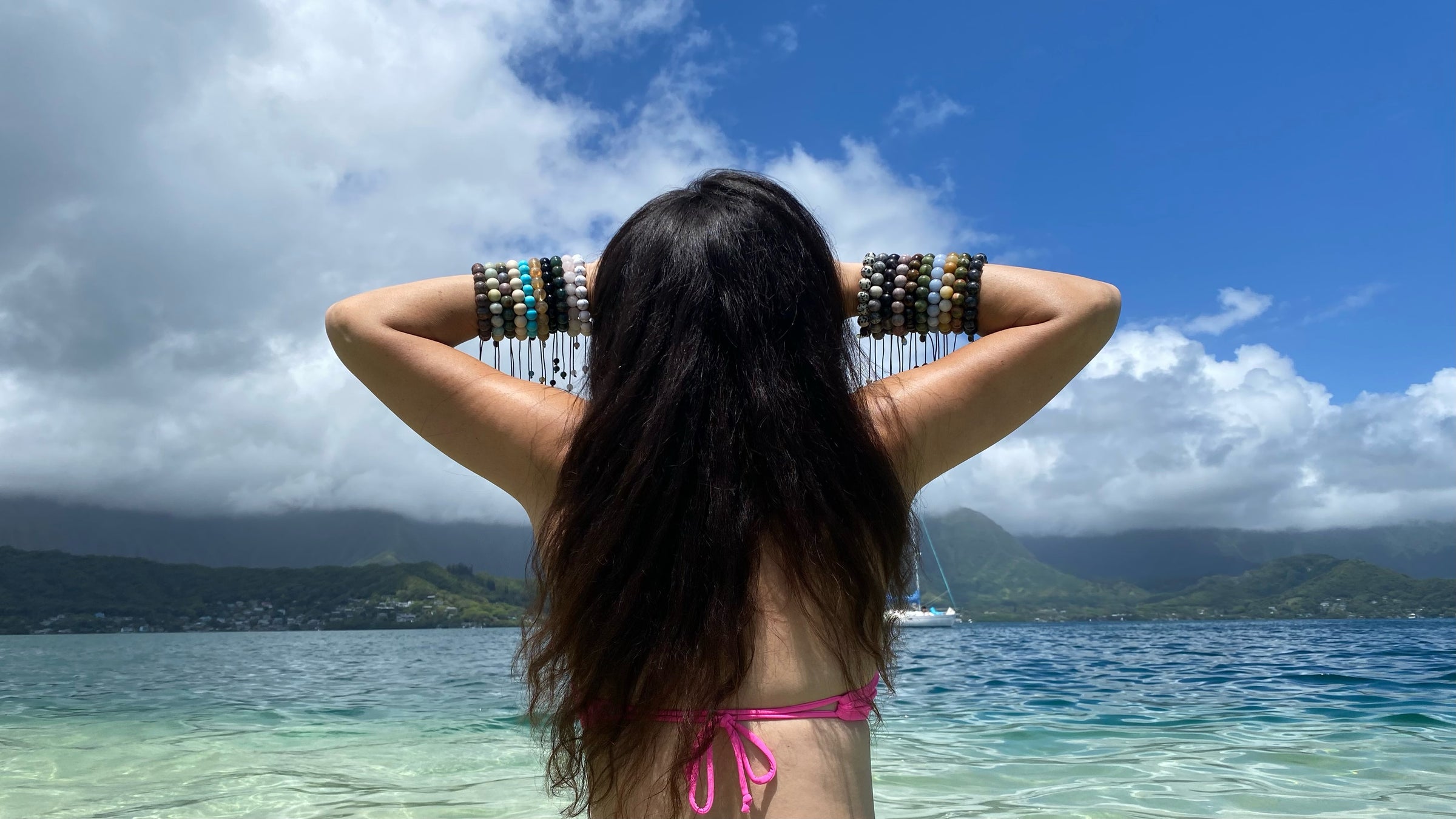 Close-up of the back of a woman with long black hair wearing a pink swimsuit top with ten large bead bracelets on each wrist and holding her hands to the front of her face with arms bent outwards, in the background there's a blue sky, stormy clouds, green mountains, the ocean water in shades of blue and green and a sailboat